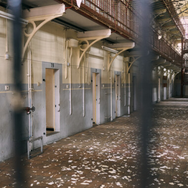 The image shows the interior of an older prison building: a long corridor of cells with several numbered doors. The floor is littered with flaking paint or plaster.